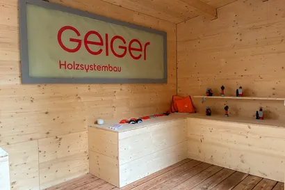 Holzstand mit einem großen "Geiger Holzsystembau"-Schild und verschiedenen kleinen Werkzeugen, die auf Regalen und Theken ausgestellt sind.