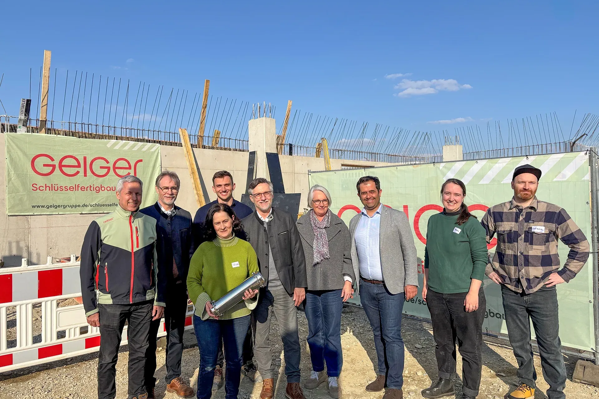 Eine Gruppe von Menschen lächelt vor einer Baustelle mit Bannern und Ausrüstung, unter einem klaren blauen Himmel.