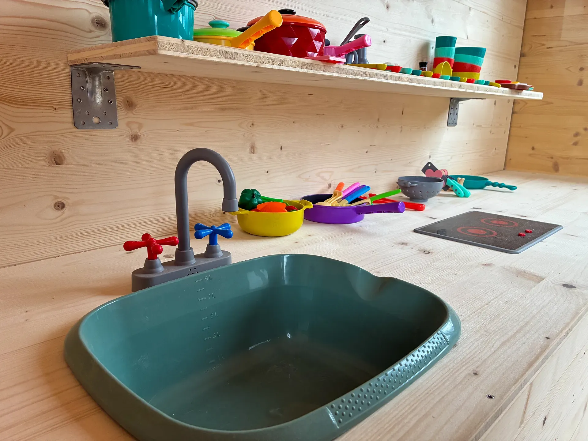 Spielküche mit einem türkisfarbenen Waschbecken, einem grauen Wasserhahn und buntem Spielküchenzubehör auf einer Holztheke und einem wandmontierten Regal.