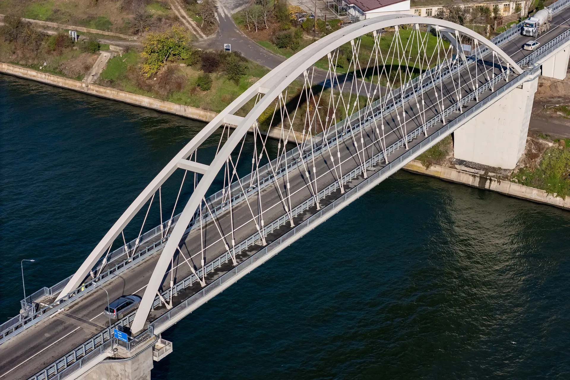 Vedere aeriană a unui pod arcuit care traversează un râu larg, cu vehicule care îl traversează și vegetație pe malurile râului.