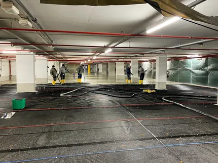 Arbeiter mit Werkzeugen und Ausrüstung reinigen einen großen Innenraum mit Rohren und schwarzem Bodenbelag unter Neonbeleuchtung.