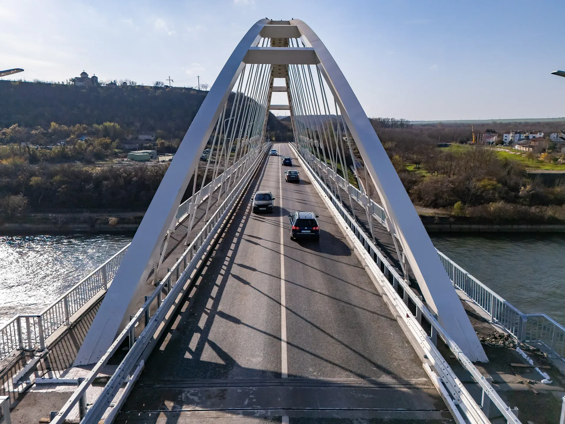 Vedere aeriană a unui pod cu suporturi arcuite peste un râu, pe care circulă vehicule într-o zi însorită, înconjurat de dealuri și vegetație rară.