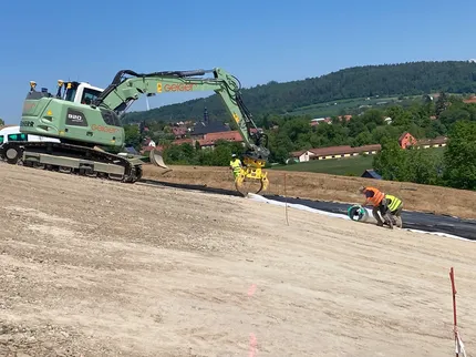 Im Zentrum ein grüner Bagger der Geiger Gruppe, während zwei Mitarbeitende eine Abdeckung den Hang hoch rollen.