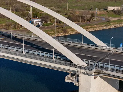 Vedere aeriană a unui pod arcuat modern peste un râu, cu drumuri asfaltate și peisaj verde în fundal.