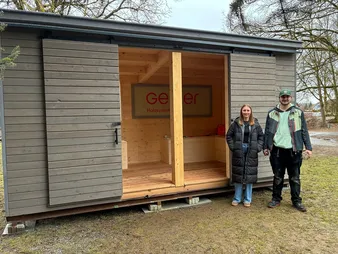 Zwei Personen stehen neben einer offenen Holzhütte mit Schiebetüren. Innen hängt ein Schild mit der Aufschrift 'Geier Holzsysteme'. Im Hintergrund stehen Bäume.