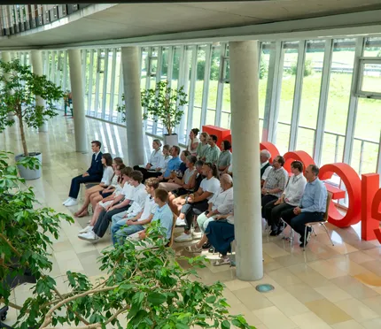 Eine Gruppe von Menschen sitzt in einem modernen, sonnenbeschienenen Flur mit bodentiefen Fenstern und großen roten Buchstaben im Hintergrund, umgeben von Grünpflanzen.