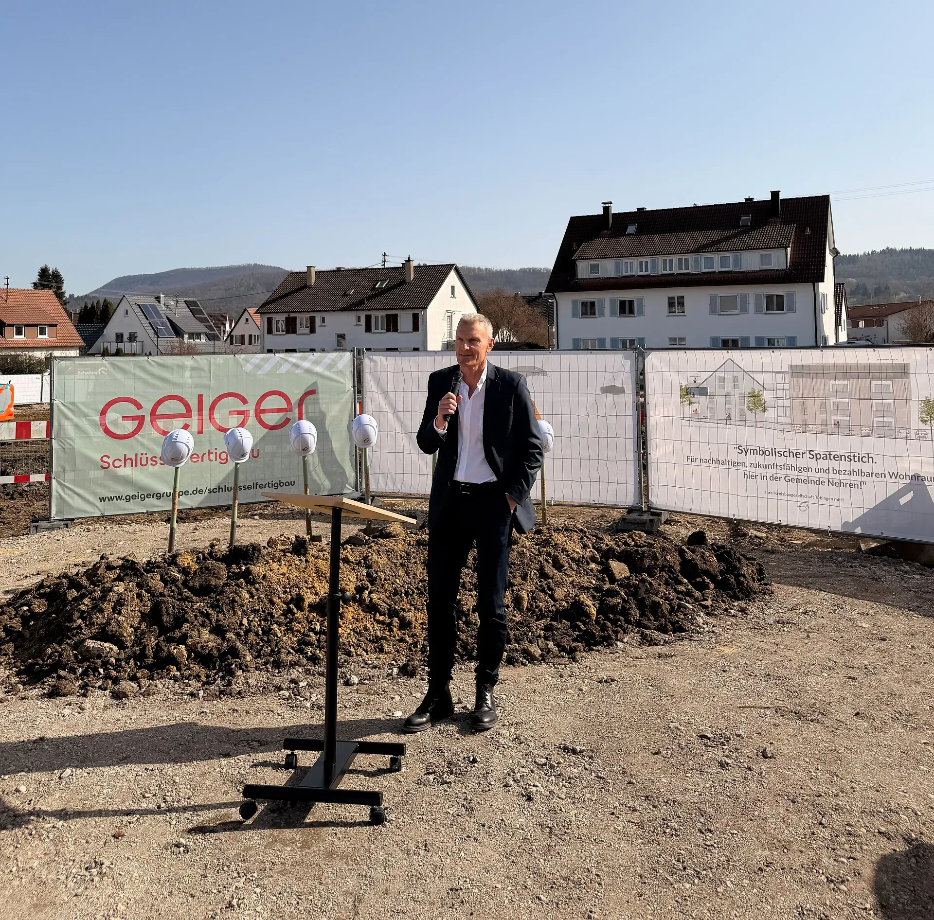Ein Mann spricht auf einer Baustelle mit einem Mikrofon und steht in der Nähe eines Podiums. Im Hintergrund sind Häuser und Banner sichtbar.