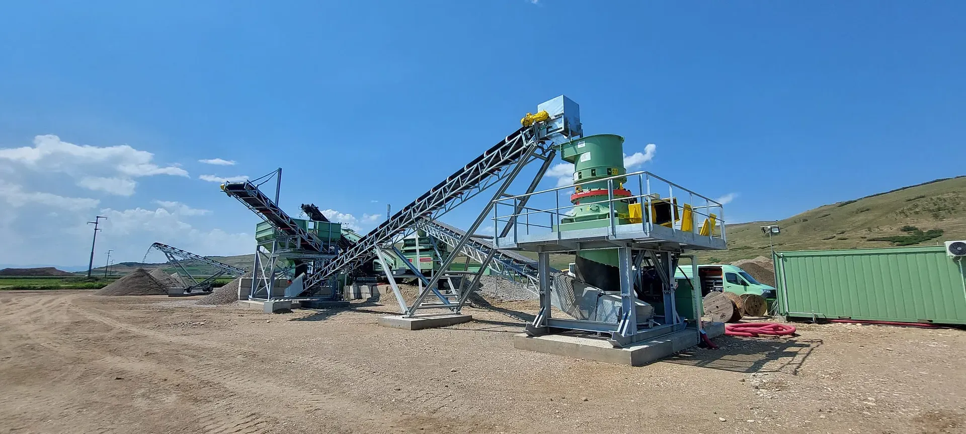 Șantier industrial în aer liber cu utilaje grele, inclusiv benzi transportoare și unități de depozitare, pe fundalul unui cer albastru senin cu dealuri îndepărtate.