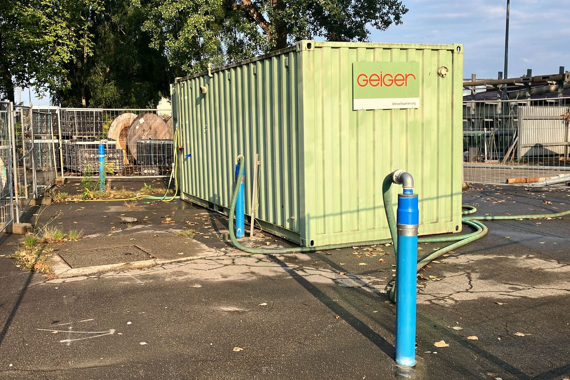 Ein grüner Schiffscontainer mit angeschlossenen Schläuchen, umgeben von Zäunen und Baumaterialien, an einem sonnigen Tag unter einem Baum.