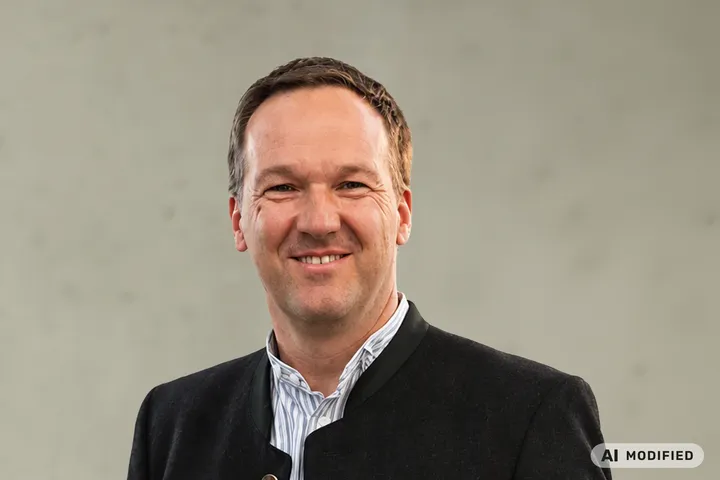 Lächelnder Mann in dunkler Trachtenjacke über einem gestreiften Hemd und Jeans, vor hellem, strukturiertem Hintergrund stehend.