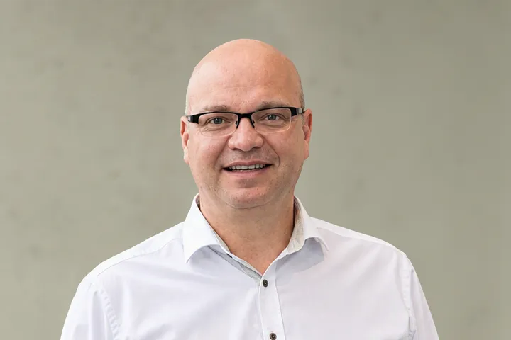 Lächelnder Mann mit Brille und Bart, der ein blau-weiß kariertes Hemd trägt, vor schlichtem Hintergrund stehend.