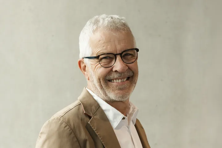 Lächelnder älterer Mann mit weißem Haar und Bart, der eine Brille, ein beigefarbenes Sakko und ein weißes Hemd trägt, vor hellgrauem Hintergrund stehend.