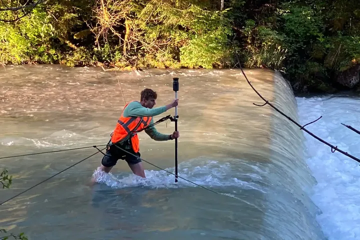 Person in Schutzausrüstung durchquert einen Fluss nahe eines Wasserfalls, hält eine Messstange und sichert sich mit Seilen, umgeben von Bäumen.