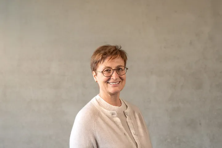 Lächelnde Person mit kurzen Haaren und Brille, die einen hellen Pullover trägt und vor einem schlichten beigen Hintergrund steht.