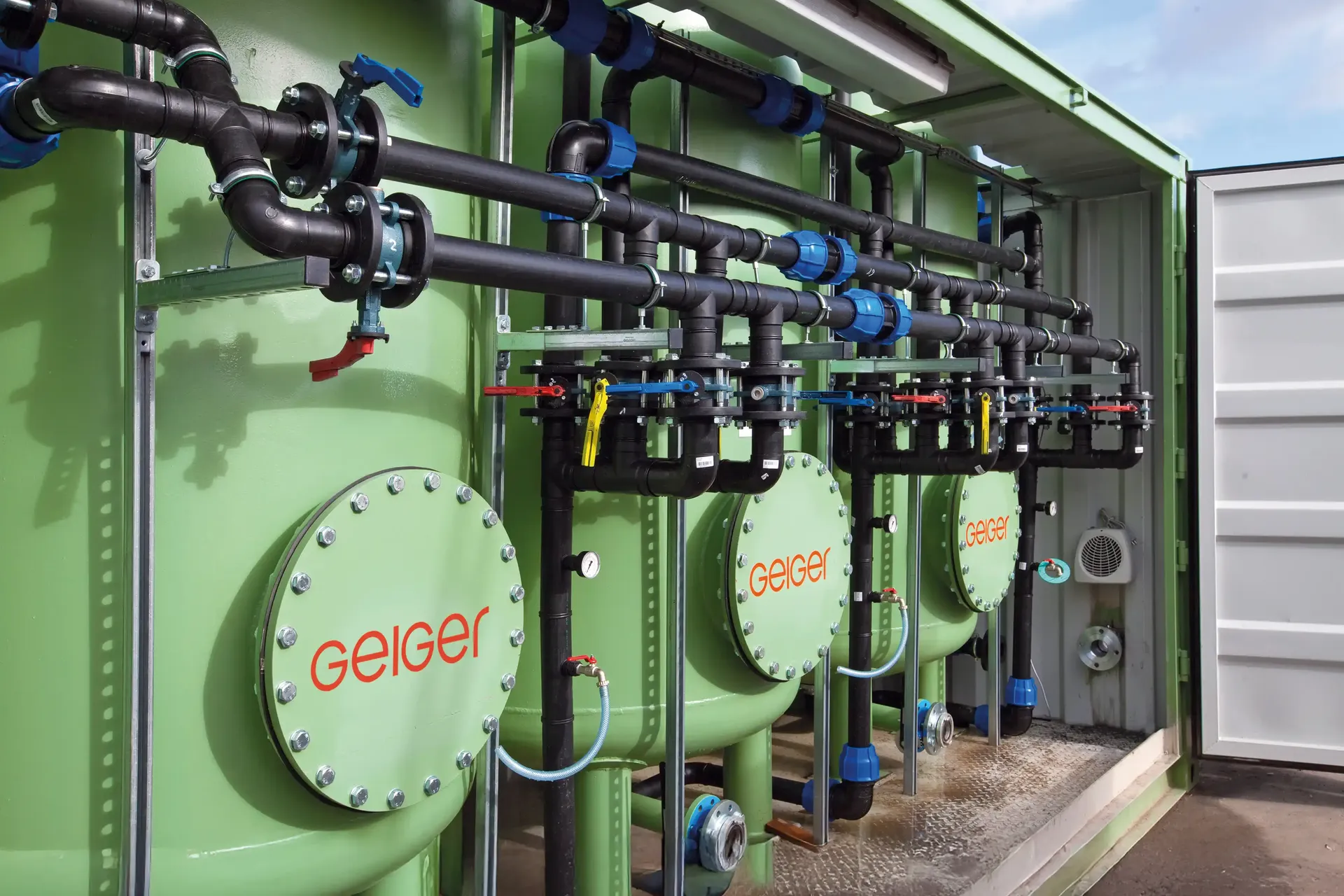Grüne Industrietanks mit schwarzen Rohrleitungen und Ventilen in einem Metallcontainer, beschriftet mit ‚geiger‘.