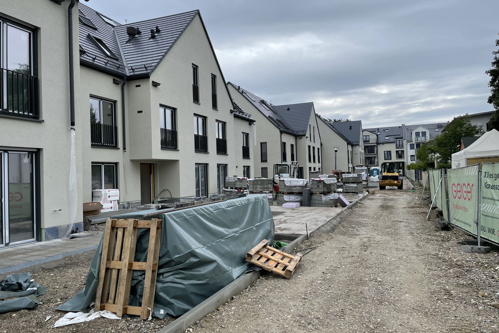 Baustelle mit modernen Stadthäusern, Baumaterialien und Geräten auf einer unbefestigten Straße unter bewölktem Himmel.