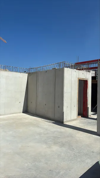 Teilweise errichtetes Gebäude mit Betonwänden und Betonboden unter einem klaren blauen Himmel. Sichtbare Bewehrungsstäbe aus Stahl sowie ein vorbereiteter Türdurchgang geben Einblick in den aktuellen Baufortschritt des Rohbaus.