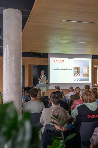 Antwort wird generiert Copilot said: Eine Person hält während einer Preisverleihung eine Rede auf der Bühne, während das Publikum aufmerksam zuhört und im Hintergrund eine Präsentationsfolie auf einer Leinwand angezeigt wird.