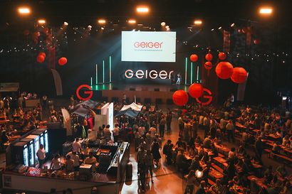 Eine große Indoor-Veranstaltung mit Publikum, Bühne und roter Dekoration. Über der Bühne zeigt ein Bildschirm „Geiger“. Stände und Lichter sind sichtbar.