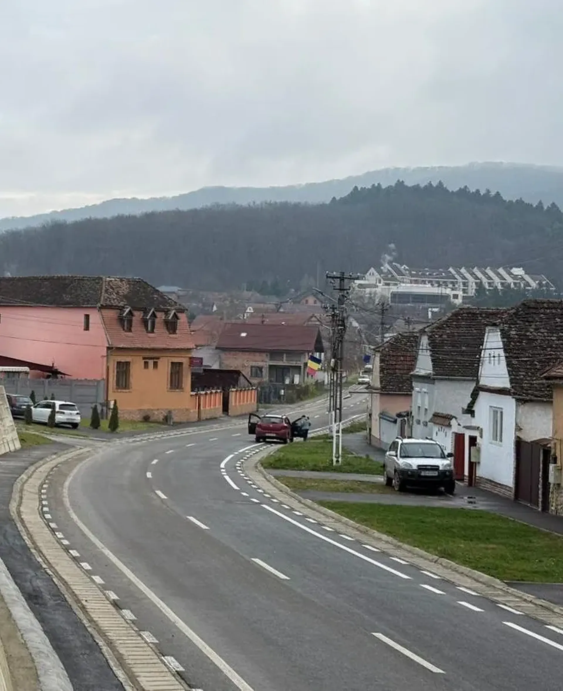 Un drum curbat mărginit de case într-un oraș mic, având în fundal dealuri domoale sub un cer înnorat.