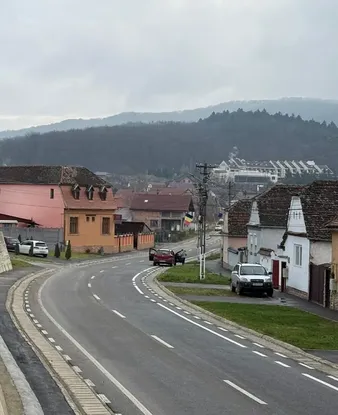 Un drum curbat mărginit de case într-un oraș mic, având în fundal dealuri domoale sub un cer înnorat.