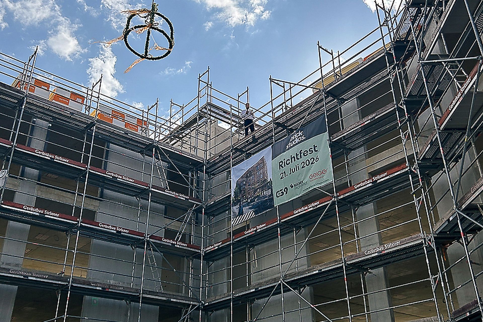 Baustelle mit einem eingerüsteten Gebäude und einem von einem Kran angehobenen Richtkranz. Zwei Männer stehen an Tischen und sprechen zu einer kleinen Gruppe von Gästen. Ein Plakat weist auf das Richtfest am 21. Juli 2026 hin.