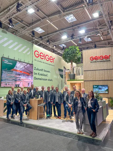 Eine Gruppe von Personen in passenden Jacken posiert an einem modernen Messestand mit Unternehmensbranding und Ausstellungsflächen.