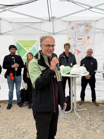 Ein Mann in einer grün-schwarzen Jacke hält ein Mikrofon und spricht unter einem Zelt zu einem Publikum. Hinter ihm liegen Bauhelme auf einem Tisch.