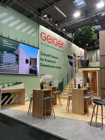 Messestand mit grünen Wänden, Barhockern, Holztischen, einer Treppe und einem großen digitalen Bildschirm, auf dem ein Architekturprojekt angezeigt wird.