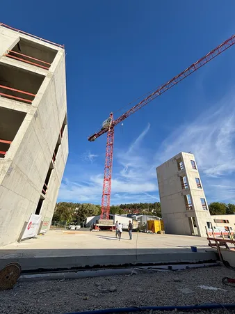Baustelle mit zwei Personen, die unter einem großen roten Kran gehen, umgeben von teilweise gebauten Betongebäuden unter klarem blauem Himmel.