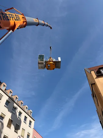 Ein Kran hebt Baumaterialien hoch über die Gebäude, unter einem klaren blauen Himmel.