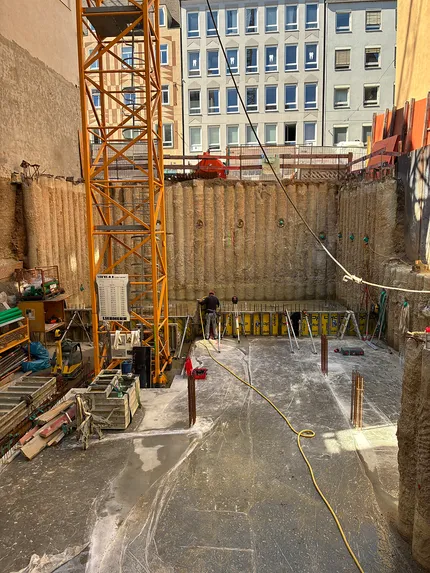 Baustelle mit einer tiefen Baugrube, einem gelben Kran und einem Arbeiter, der die Ausrüstung anpasst. Im Hintergrund Gebäude, die von Sonnenlicht beleuchtet werden.