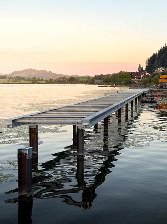 Ein Metallsteg ragt bei Sonnenuntergang über ruhiges Wasser hinaus, mit Hügeln und Gebäuden im Hintergrund.