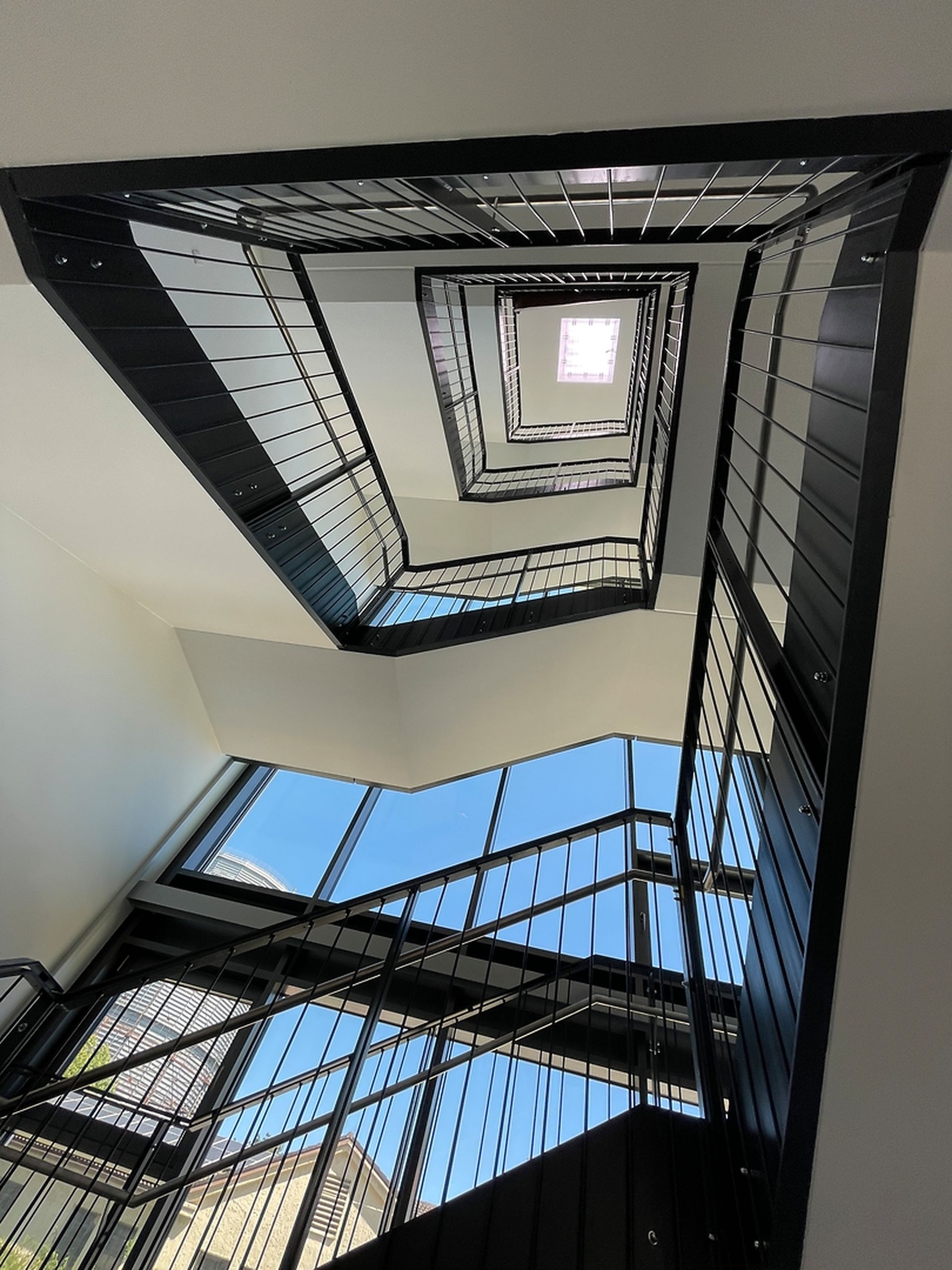 Blick nach oben auf eine moderne, schwarze Metalltreppe mit geometrischen Linien und einem Oberlicht an der Spitze, eingerahmt von großen Fenstern.