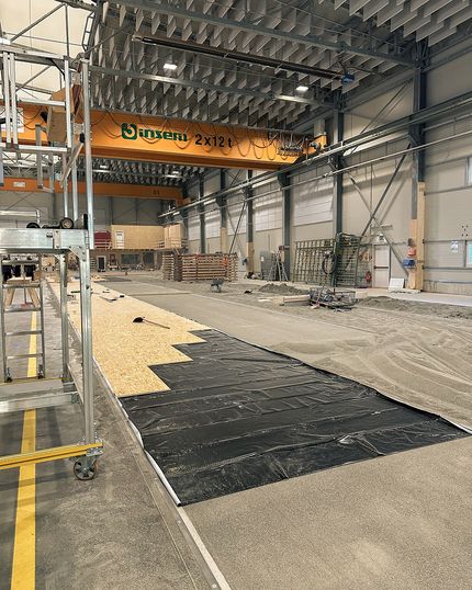 Fertigungsstraße bei Geiger Holzsystembau mit schwarzen Kunststofffolien, Holzplatten und einer sandigen Unterlage. Darüber ist ein großer Hallenkran zu sehen.