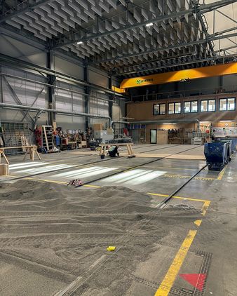 Innenansicht einer der Werkhalle bei Geiger Holzsystembau mit moderner Ausstattung. Im Vordergrund ist ein Haufen von Kies oder Sand zu sehen, während im Hintergrund Maschinen und Arbeitsgeräte neben einer Holzplattform angeordnet sind. Eine gelbe Hallenkranstruktur ist oberhalb erkennbar.