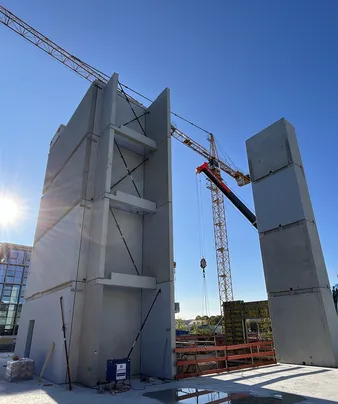 Baustelle mit großen aufgestellten Betonplatten, umgeben von Kränen unter klarem blauem Himmel. Sonnenlicht wirft Schatten auf die Baustelle.