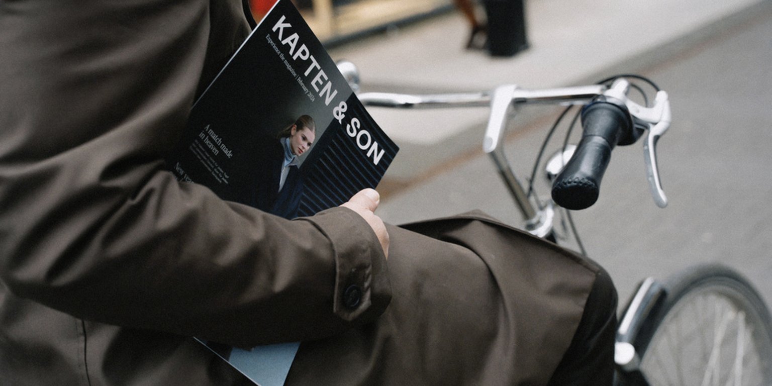 Een persoon zit naast een fiets en houdt een Kapten & Son magazine vast. Ze dragen een bruine jas.