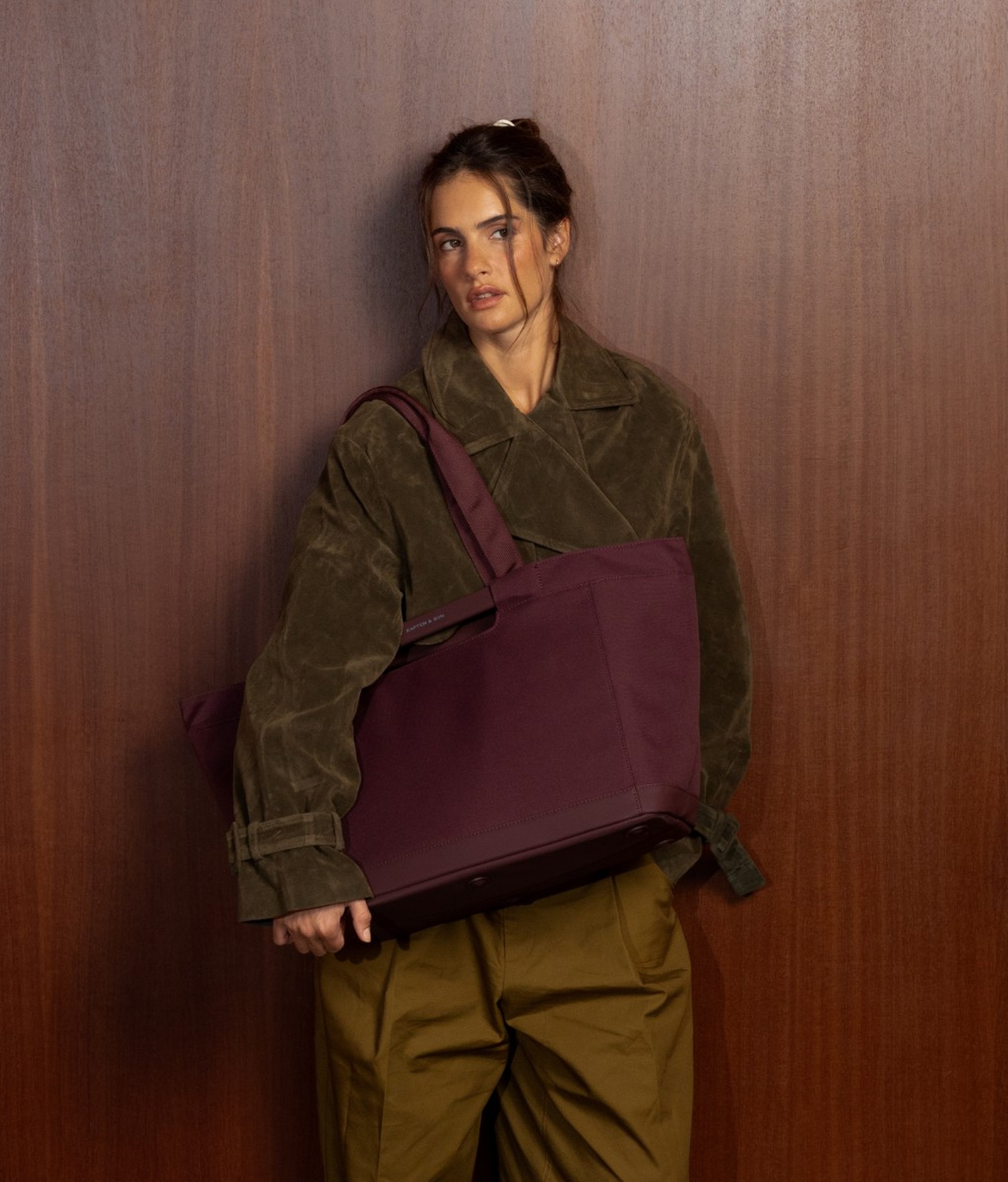 Jeune femme aux cheveux foncés en chignon lâche appuyée contre un mur lambrissé en bois foncé, portant une veste en daim vert olive et un pantalon large vert olive foncé, avec un grand sac tote Kapten & Son Vaasa bordeaux avec poignées supérieures sur l'épaule.