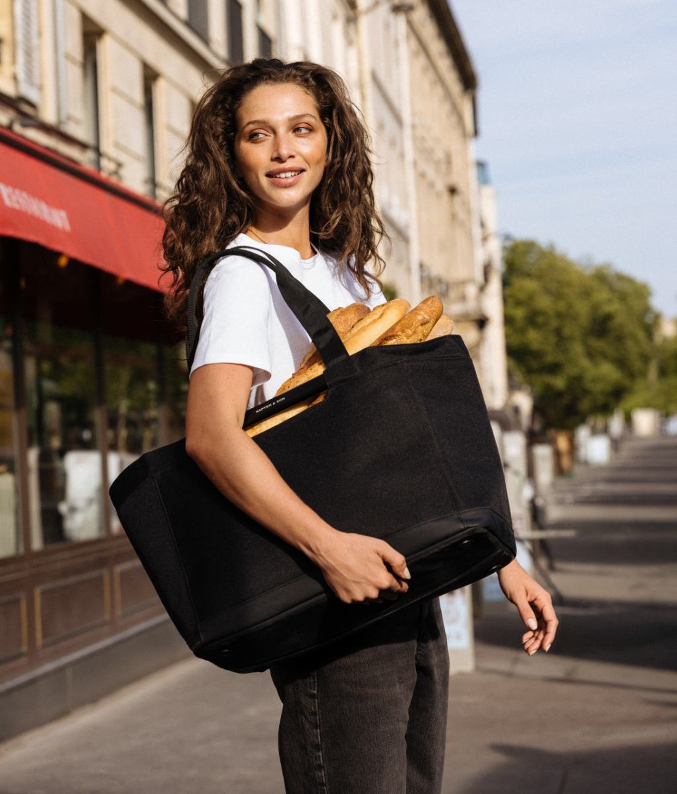 Lächelnde junge Frau mit langen lockigen Haaren trägt ein weißes T-Shirt und dunkle Jeans, hat eine große schwarze Kapten & Son Shopping-Tote gefüllt mit Baguettes über der Schulter, geht auf einer sonnigen Pariser Straße mit einem Restaurant-Markisenzeichen im Hintergrund.