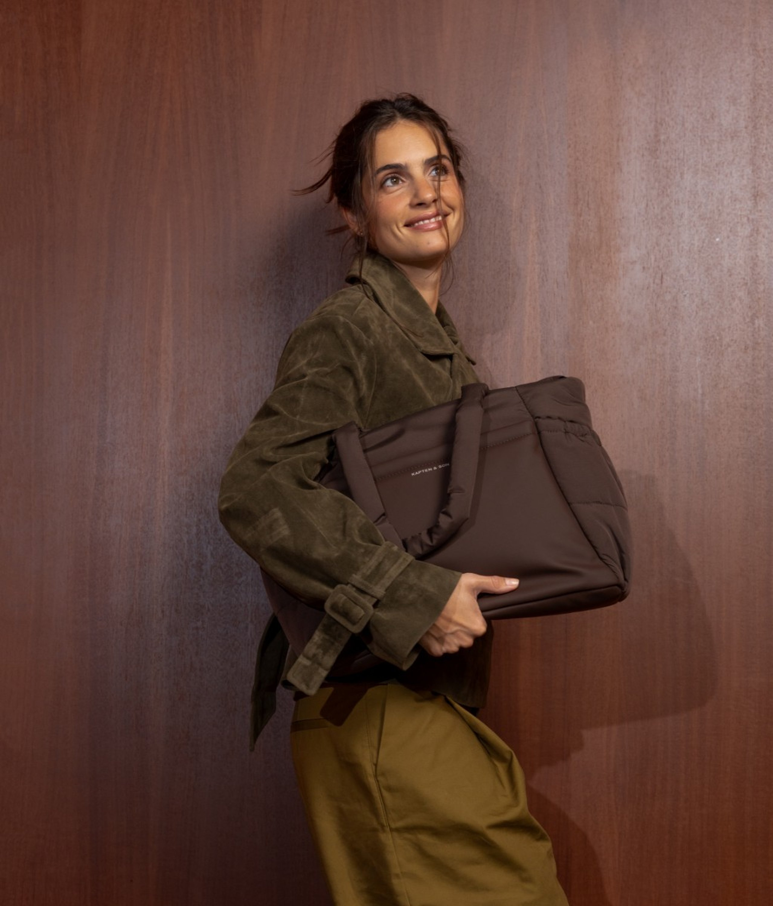 Jeune femme souriante aux cheveux foncés en chignon lâche appuyée contre un mur lambrissé en bois foncé, portant une veste en daim vert olive et un pantalon large vert olive foncé, tenant un grand sac tote matelassé Kapten & Son Hellvi Cloud couleur chocolat avec poignées supérieures dans ses bras.