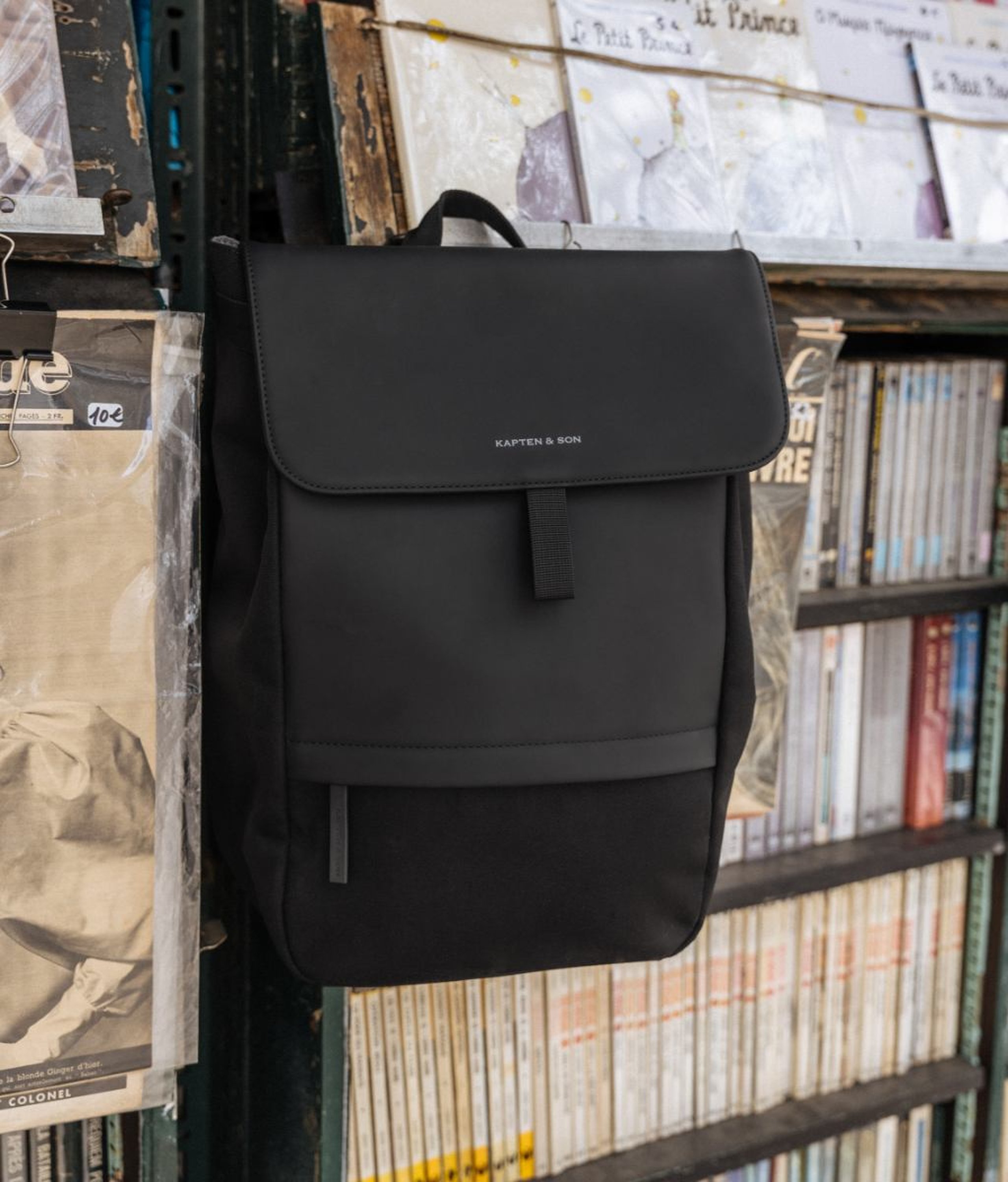Black Kapten & Son flap backpack with a top handle, a magnetic closure flap and a front zip pocket, hanging on a Parisian riverside bookstall shelf surrounded by books, magazines and vintage prints.