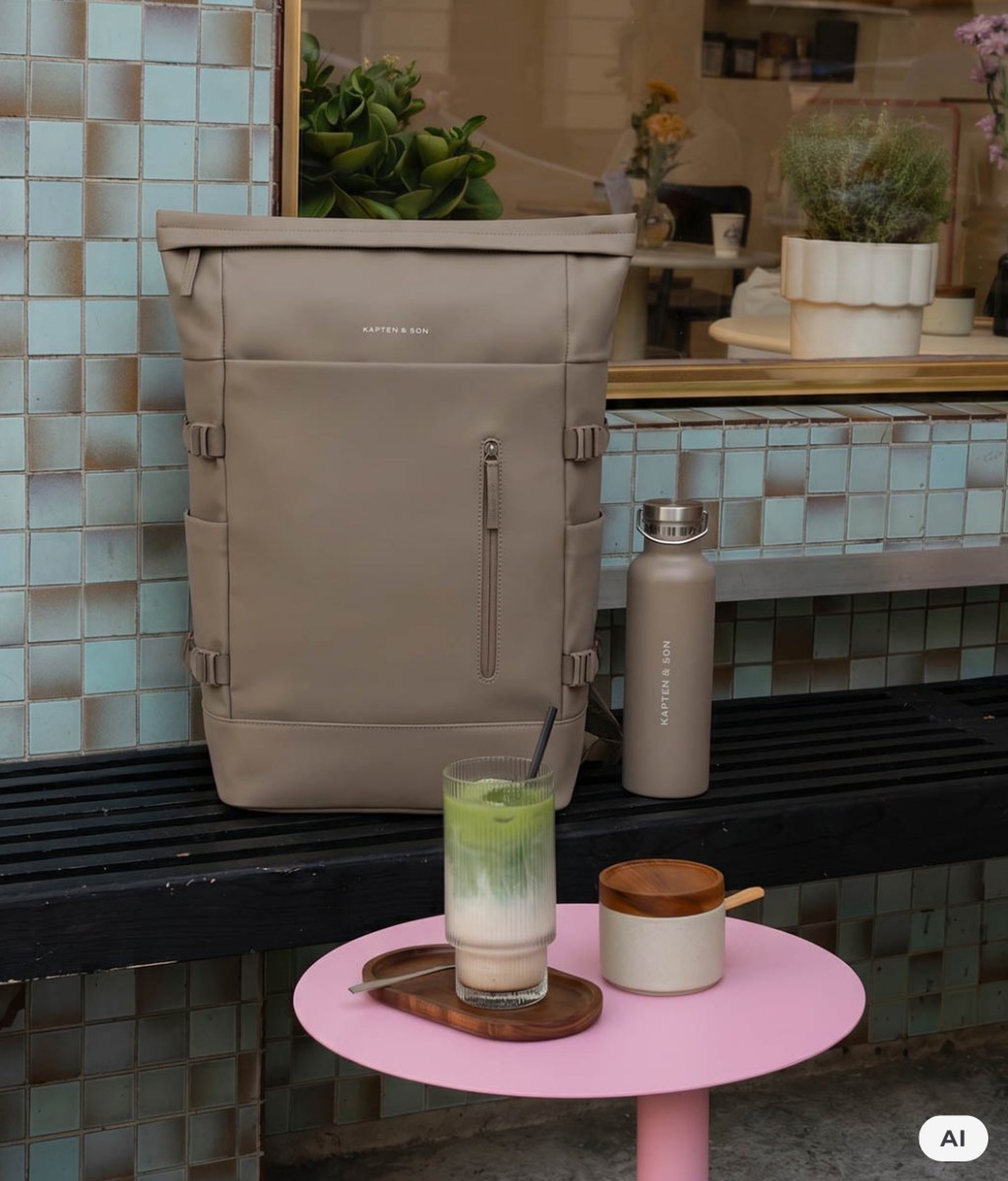 AI Generated: Ein taupefarbener Kapten & Son Rolltop-Rucksack und eine passende Kapten & Son Trinkflasche stehen auf einer Bank vor einem Café, daneben ein Matcha-Getränk auf einem rosa Tisch.