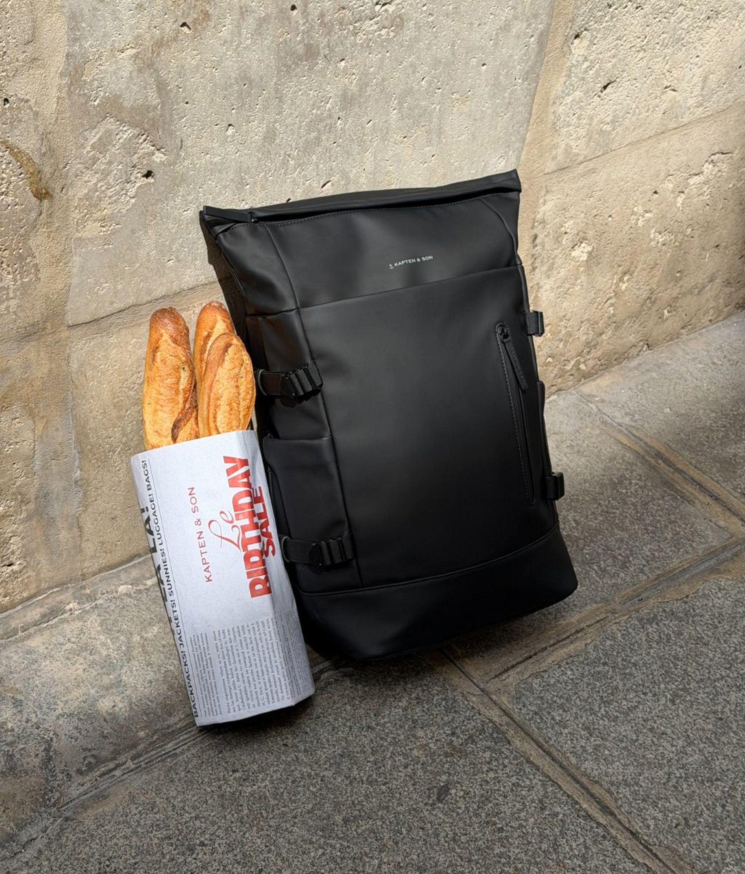 Black Kapten & Son roll-top backpack with side buckles and a front zip pocket, leaning against a stone wall next to a "Le Birthday Sale – Kapten & Son" newspaper wrapping filled with baguettes, on a stone pavement.