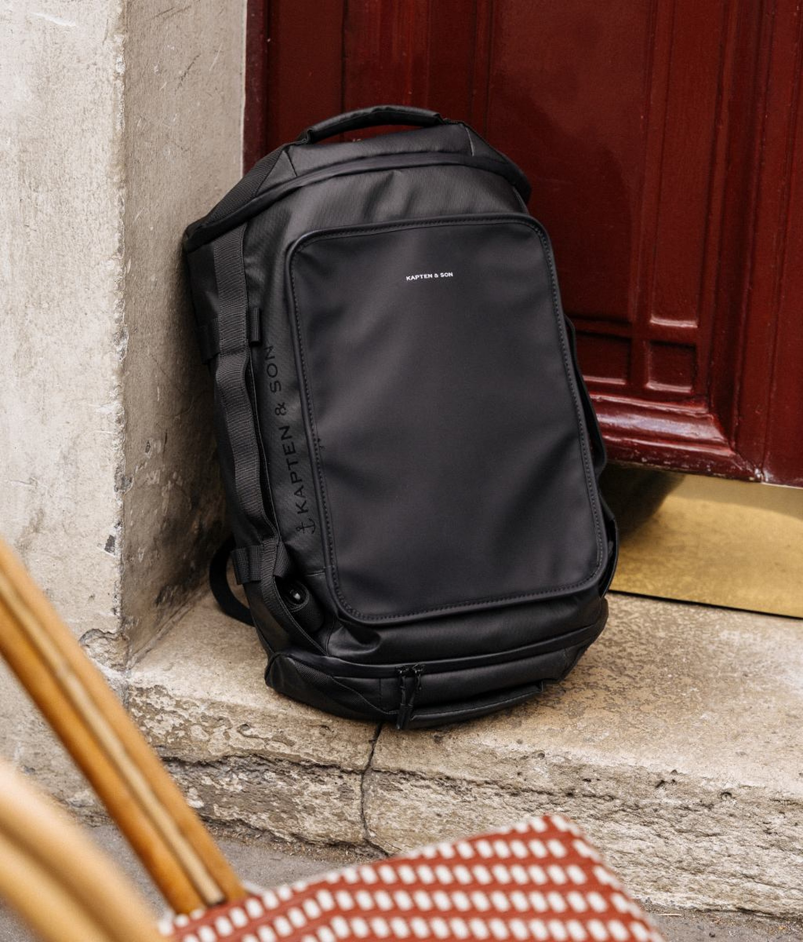 Large black Kapten & Son travel backpack with a top handle, structured front pocket, side straps and multiple compartments, leaning against a stone wall next to a dark red Parisian entrance door, with a red wicker chair visible in the foreground.