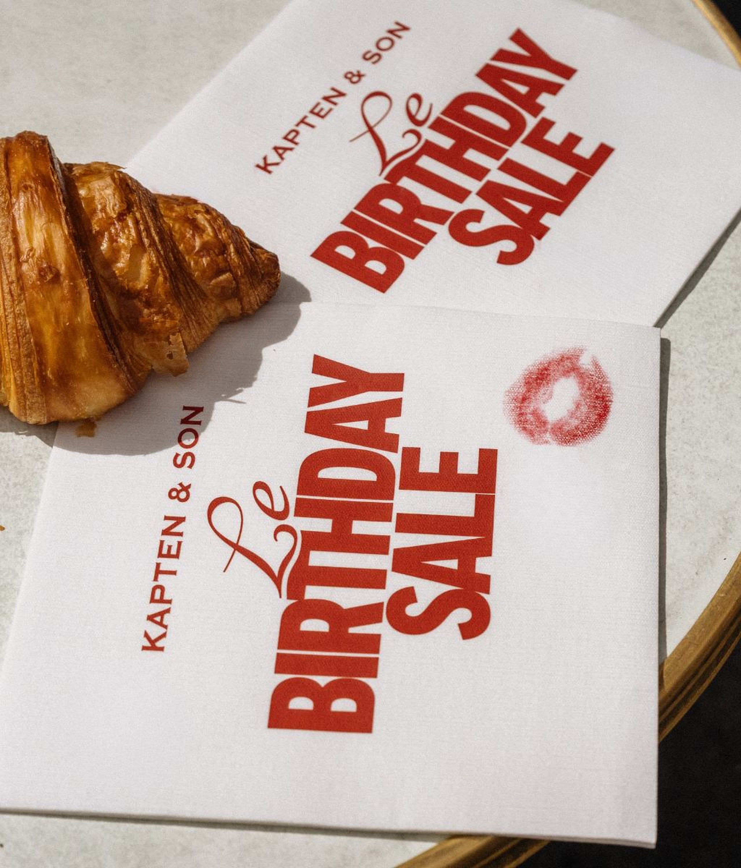 Zwei weiße Kapten & Son Papierservietten mit dem Aufdruck „Le Birthday Sale" in roten Buchstaben, auf einem Cafétisch neben einem Croissant, eine Serviette mit einem roten Lippenstift-Kussabdruck.