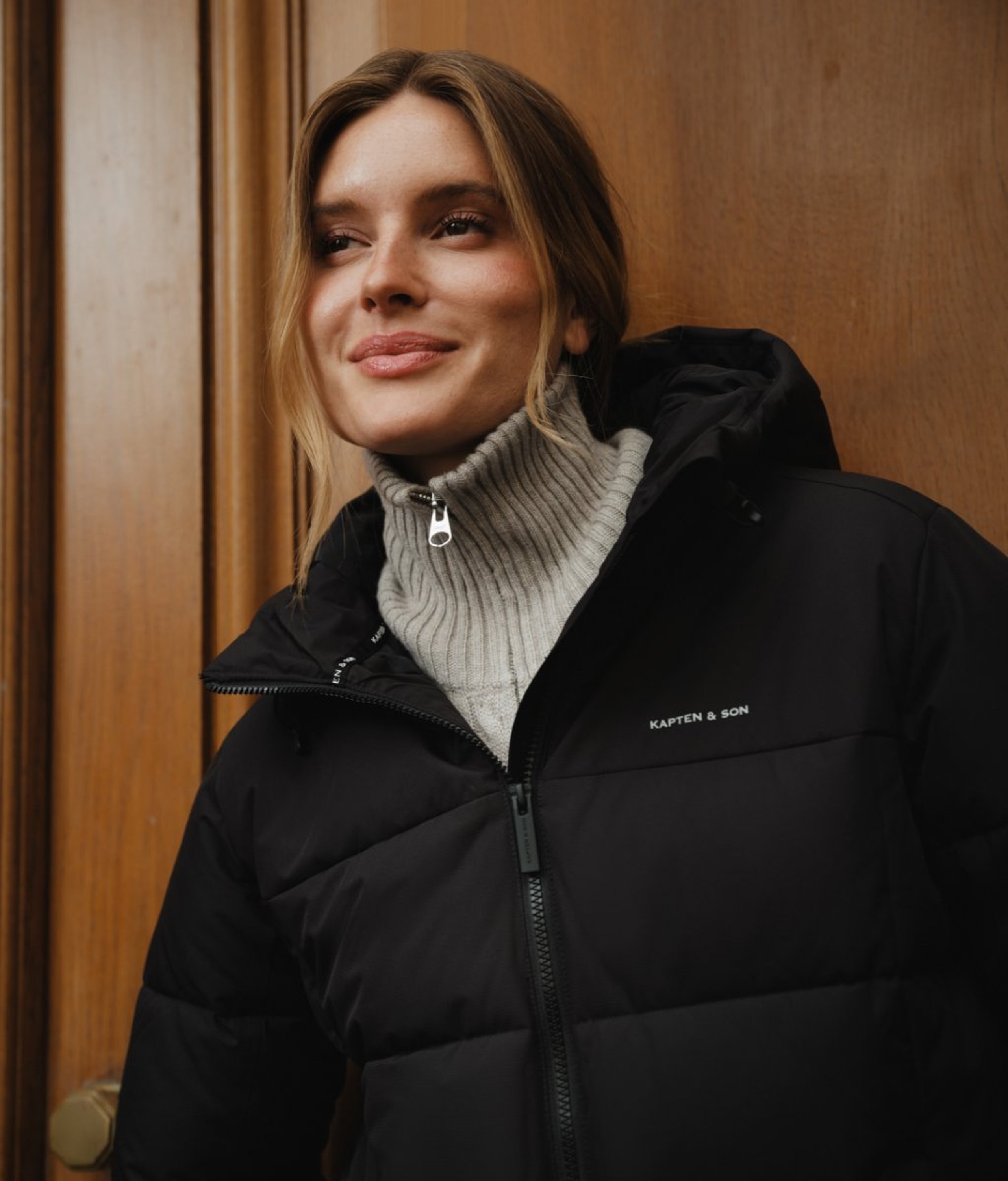 Junge Frau mit dunklem Haar sitzt seitlich in einem braunen Samtsessel mit Chrombeinen, trägt eine dunkelbraune Lederjacke über einem hellblauen Hemd und hellblauen weiten Jeans mit burgunderroten Loafern, hält eine große schokoladenbraun-hellblau gestreifte Kapten & Son Hellvi Cloud Tote Bag in den Armen.