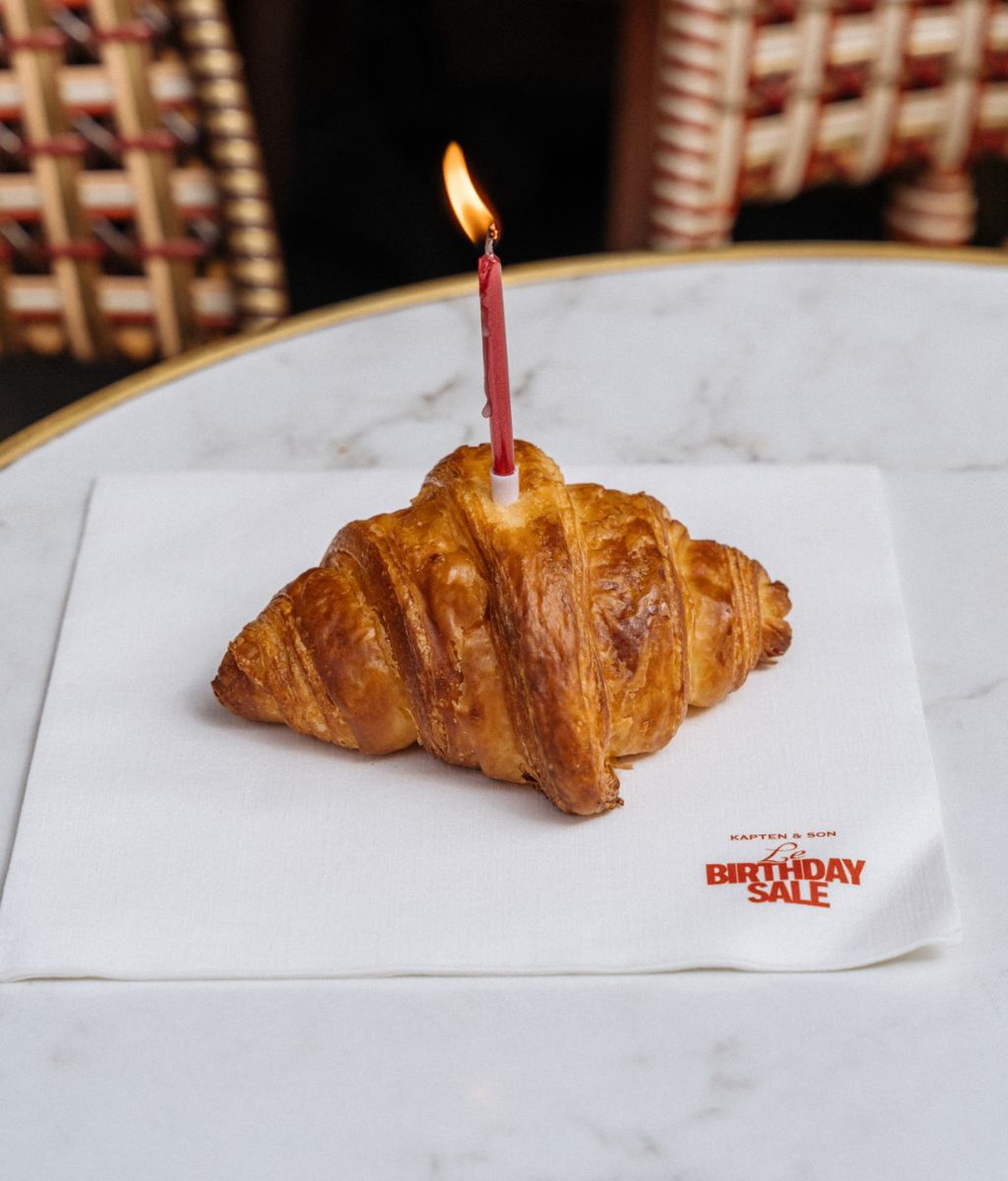 Ein goldenes Croissant mit einer brennenden roten Geburtstagskerze obendrauf, auf einer weißen Kapten & Son „Le Birthday Sale" Papierserviette auf einem Marmor-Cafétisch, mit einem roten Korbstuhl im Hintergrund.