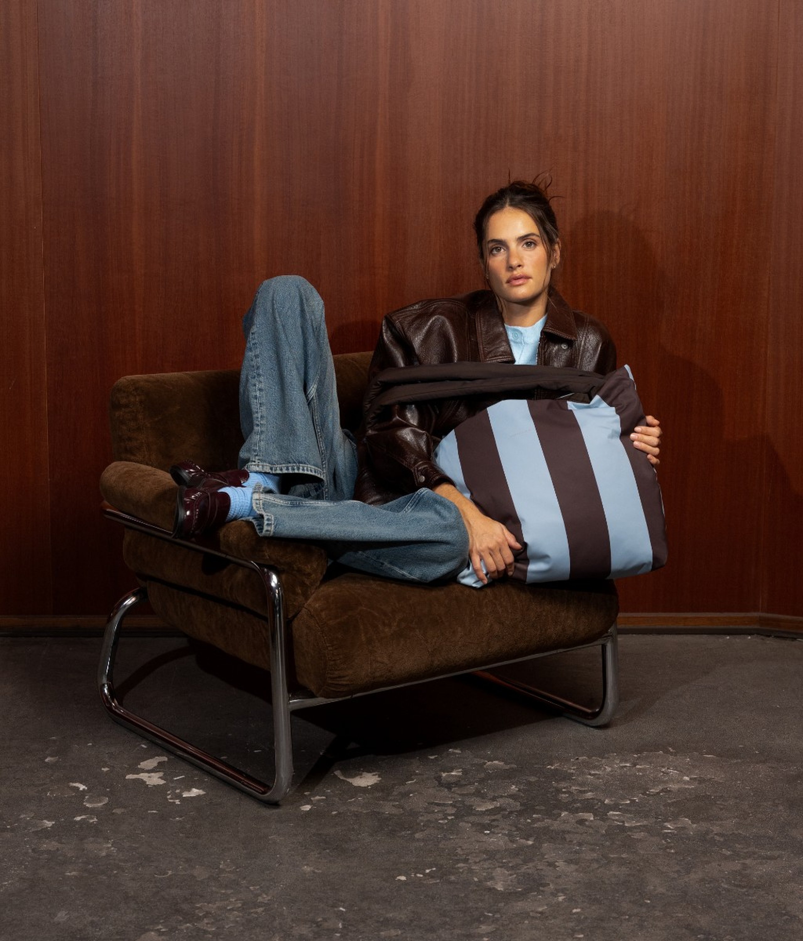 Jeune femme aux cheveux foncés assise de côté dans un fauteuil en velours marron avec des pieds chromés, portant une veste en cuir marron foncé sur une chemise bleu clair et un jean large bleu clair avec des mocassins bordeaux, tenant un grand sac tote Kapten & Son Hellvi Cloud à rayures marron chocolat et bleu clair dans ses bras.