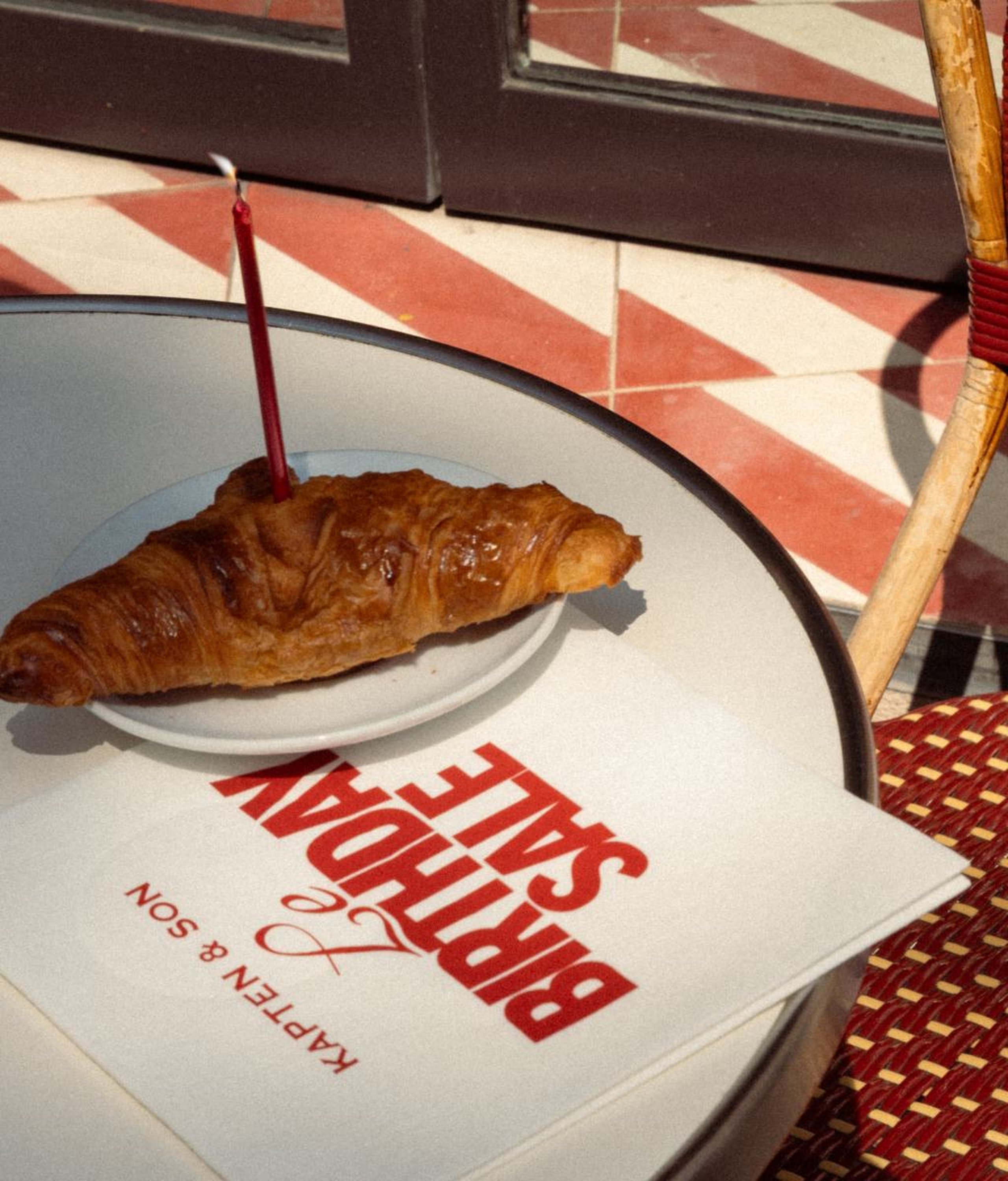 Ein Croissant mit einer unangezündeten roten Geburtstagskerze auf einem weißen Teller, auf einem Bistrotisch neben einem „Le Birthday Sale – Kapten & Son" Flyer, mit einem rot gemusterten Fliesenboden und einem Korbstuhl im Hintergrund.